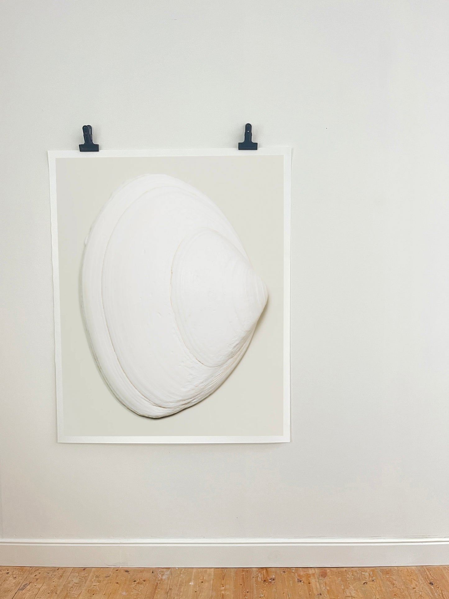 A minimalist white sculptural art piece resembling a stylized clam or shell, mounted within a white square frame on a light gray wall with two black clips at the top, above a light wood floor.