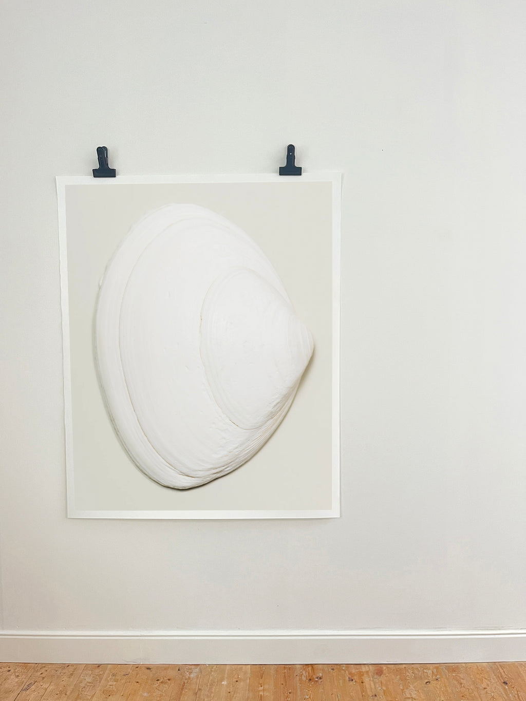 A minimalist white sculptural art piece resembling a stylized clam or shell, mounted within a white square frame on a light gray wall with two black clips at the top, above a light wood floor.