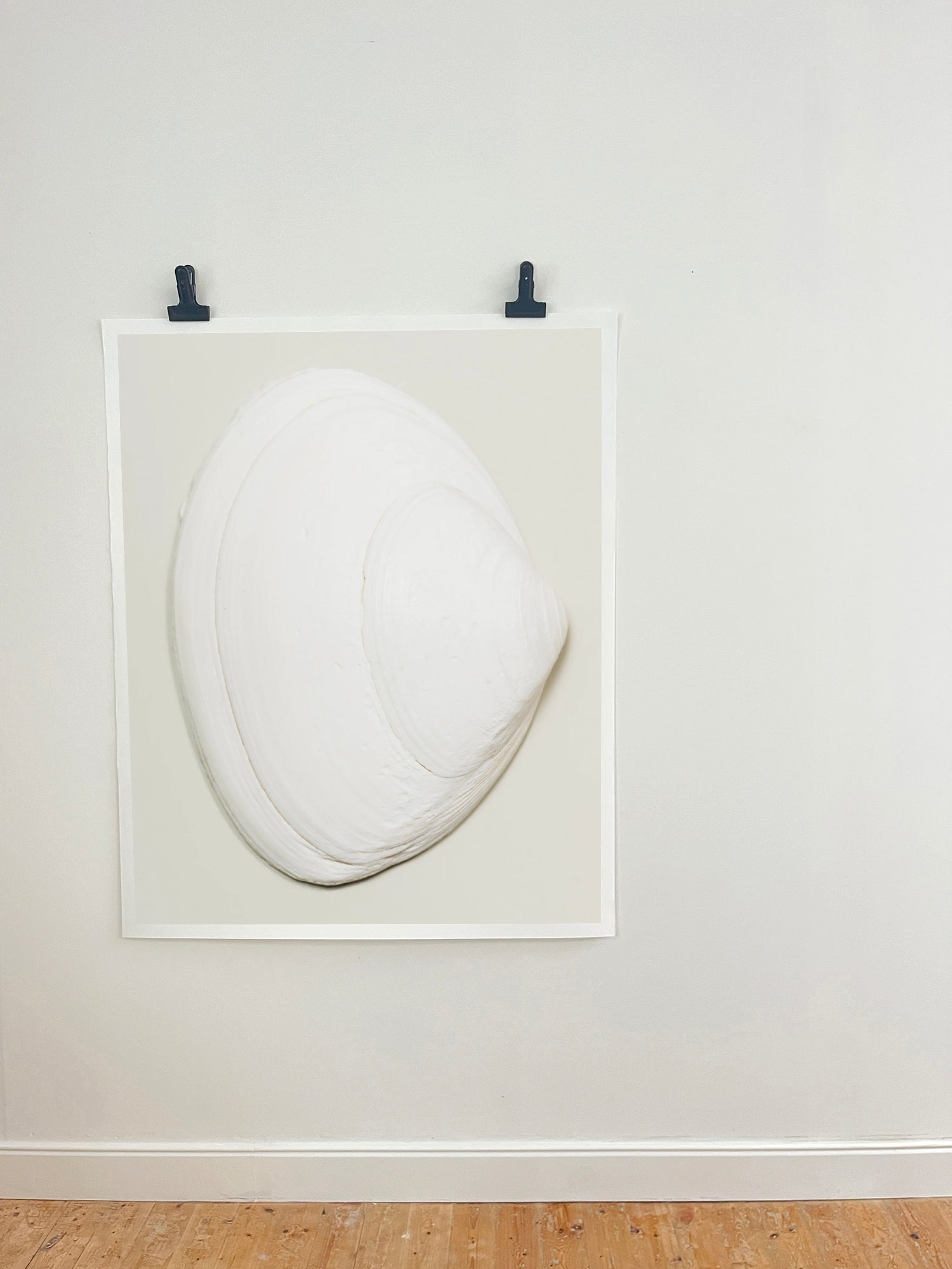 A minimalist white sculptural art piece resembling a stylized clam or shell, mounted within a white square frame on a light gray wall with two black clips at the top, above a light wood floor.