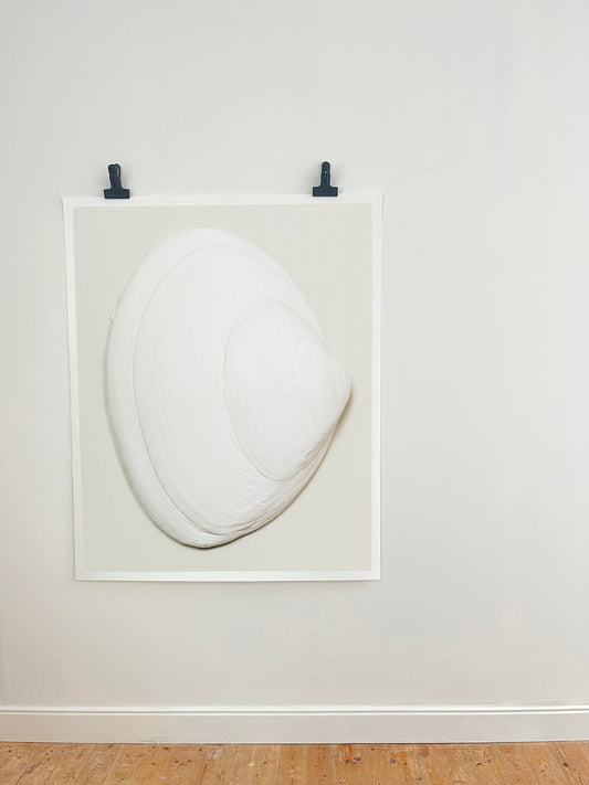 A minimalist white sculptural art piece resembling a stylized clam or shell, mounted within a white square frame on a light gray wall with two black clips at the top, above a light wood floor.